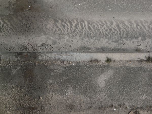 Road edge texture, with uneven grey asphalt and a cement area with a swirling discoloration, divided by a strip of similarly colored stone.